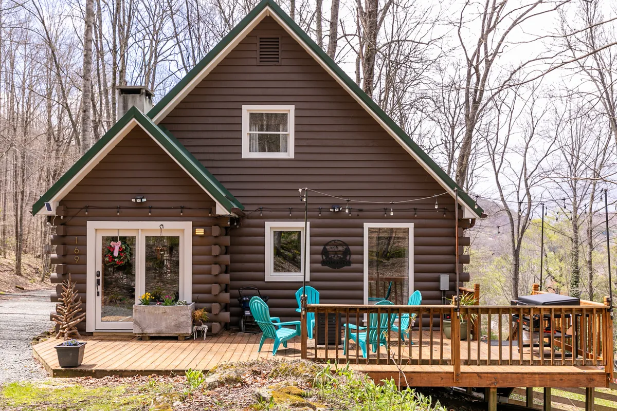 This image features the log cabin exterior with a deck furnished with chairs, large windows that reflect the outdoors, and decorative planters. The gravel path and natural surroundings add to the mountain retreat feel.