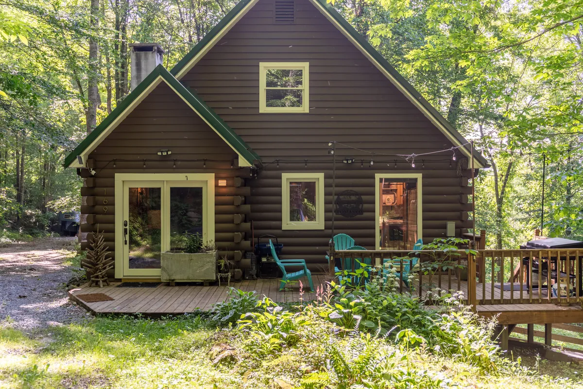 This image shows the exterior of a log cabin structure with a spacious deck area, green chairs, and surrounding greenery. The inviting deck is a perfect place for relaxing and enjoying the outdoors, with ample space for both people and pets to unwind or play in a natural setting.