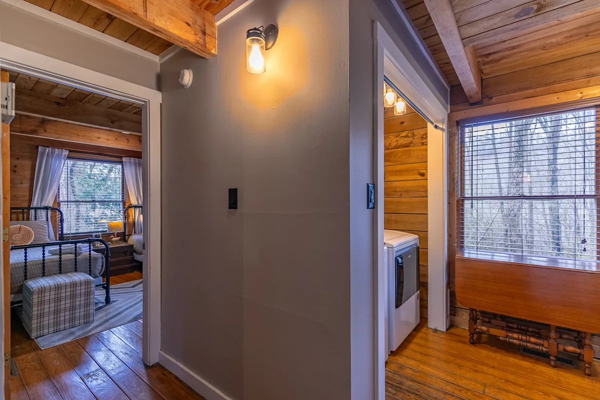 A warmly lit space featuring rustic wooden beams on the ceiling, a large window allowing natural light to pour in, a decorative light fixture, and cozy rustic furniture. The window hints at the surrounding landscape and the cabin’s placement within nature, creating a gentle bridge between the interior and the world outside.