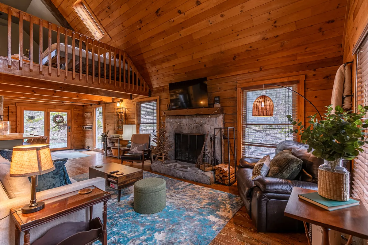 A spacious great room with a stone fireplace, comfortable sofa, decorative rug, rustic wooden beams, and expansive windows offering views of surrounding nature. The setting encourages relaxing by the fire, reading, or hosting family activities.
