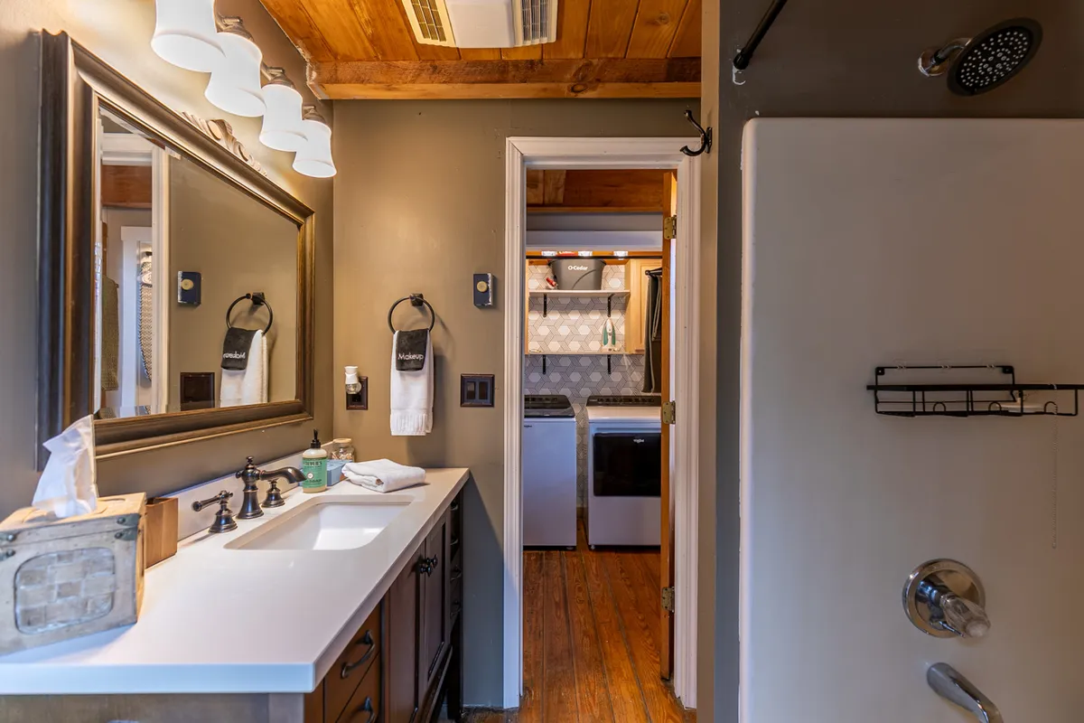A tidy bathroom with a vanity, mirror, shower, and a view toward the laundry room entrance. The space is bright and functional, offering both grooming and laundry amenities conveniently close.
