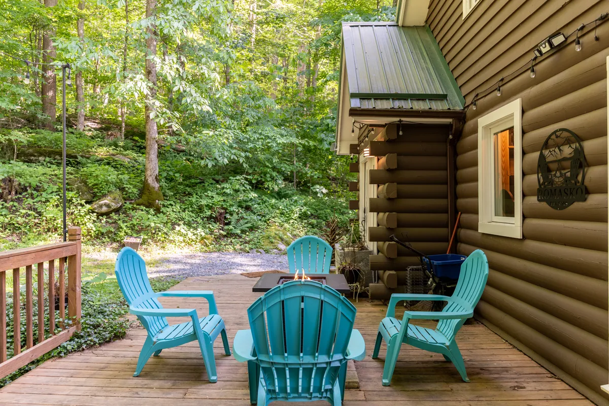 A wooden deck furnished with blue Adirondack chairs offers sweeping views of surrounding trees and greenery. The image includes part of the cabin’s exterior, emphasizing the connection between indoor comfort and outdoor exploration.