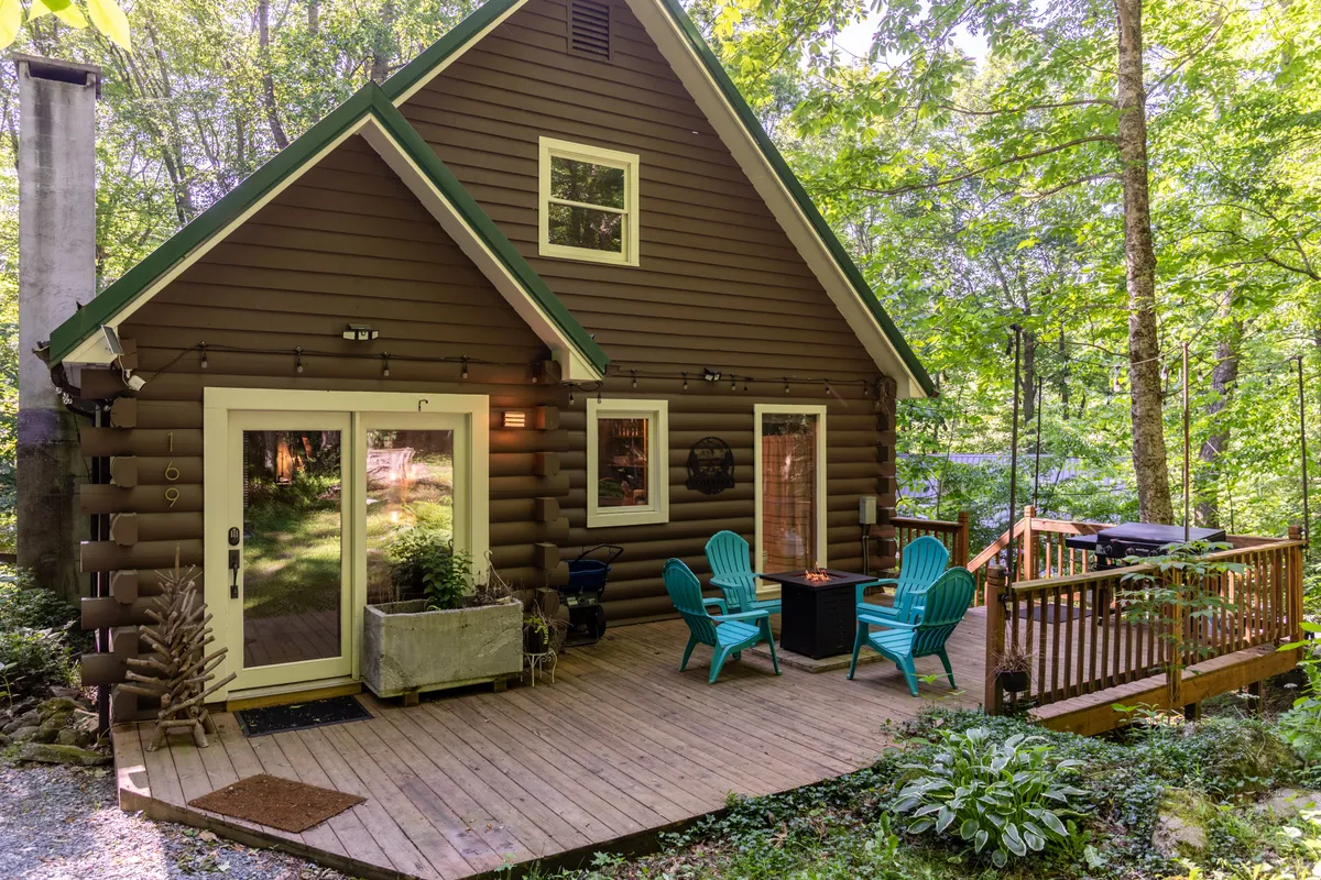 The hero image showcases a classic log cabin perched on a lush hillside, featuring a spacious deck with cozy outdoor furniture surrounded by vibrant greenery. The scene suggests tranquil mornings with sunrise views, peaceful afternoons spent relaxing outside, and the promise of stargazing under clear mountain skies.
