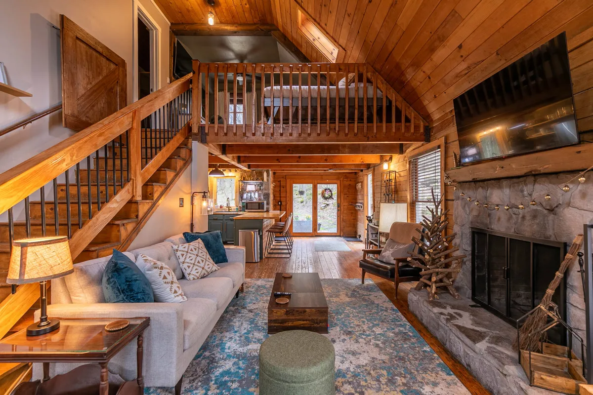 A warmly lit great room featuring a prominent stone woodburning fireplace, plush sofa, wooden stairs leading to a loft area, and a decorative tree. There’s a cozy atmosphere ideal for relaxing, socializing, and unwinding after a day outdoors.