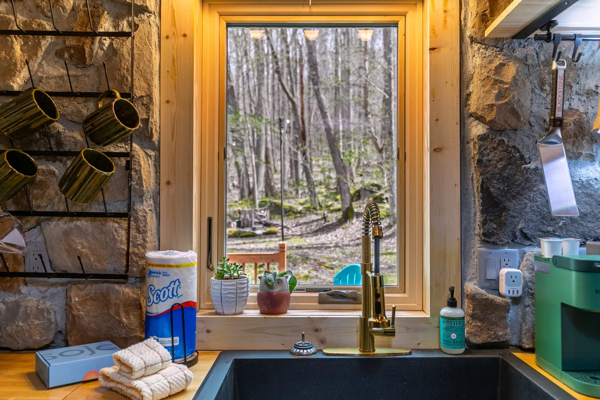 This image shows a cozy kitchen area with a kitchen sink set below a window, hanging mugs, rustic stone walls, and views of the outdoors framed by the window. The scene evokes a sense of connection to nature and hints at the changing seasons visible through the window.