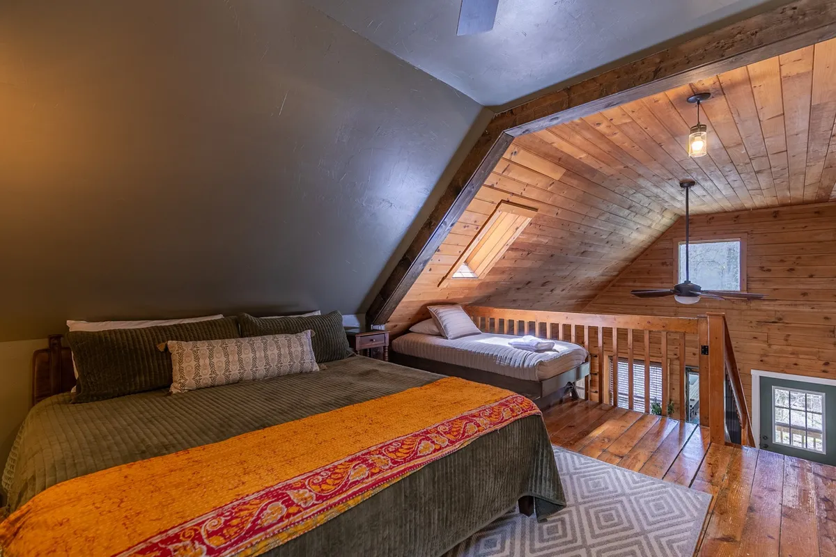 An inviting bedroom featuring a king-sized bed with a plush accent throw, wooden railing, and a sloped ceiling. The room feels spacious, serene, and bathed in soft natural light, offering a restful retreat for adults.
