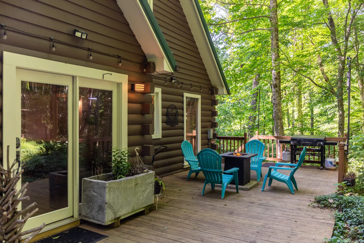 Deck seating surrounds a fire pit, with a grill and planter box adding to the amenities. The scene is casual and comfortable, with a wooded backdrop ideal for both daytime and nighttime gatherings.
