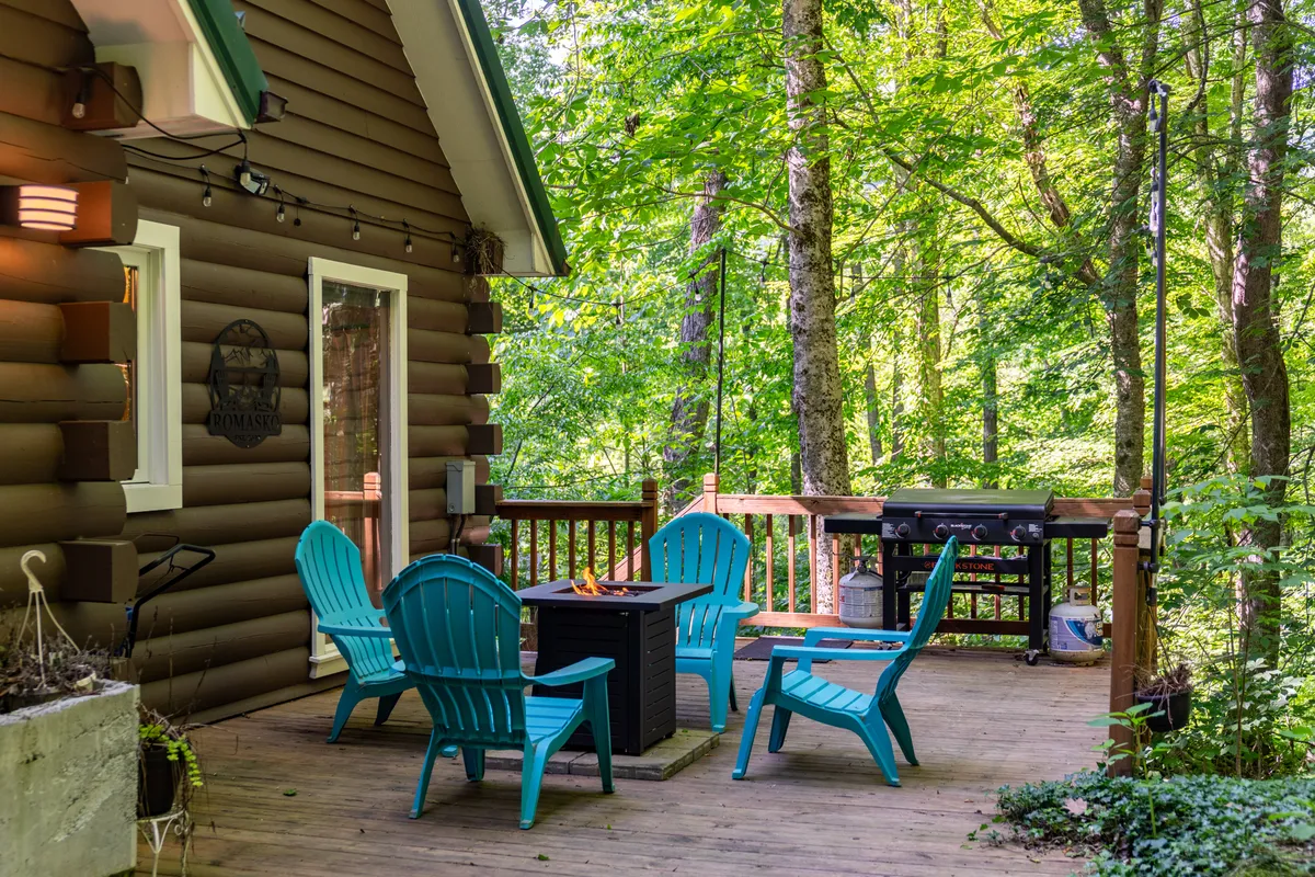 A wooden deck set amidst greenery, featuring turquoise Adirondack chairs gathered around a fire pit, along with a grill for outdoor cooking and lush natural surroundings.