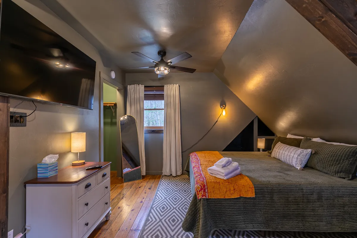 A cozy bedroom with a bed dressed in decorative throws, a television mounted on the wall, a curtained window letting in natural light, and a soft area rug. The space feels comfortable and kid-friendly, perfect for relaxing or watching TV.