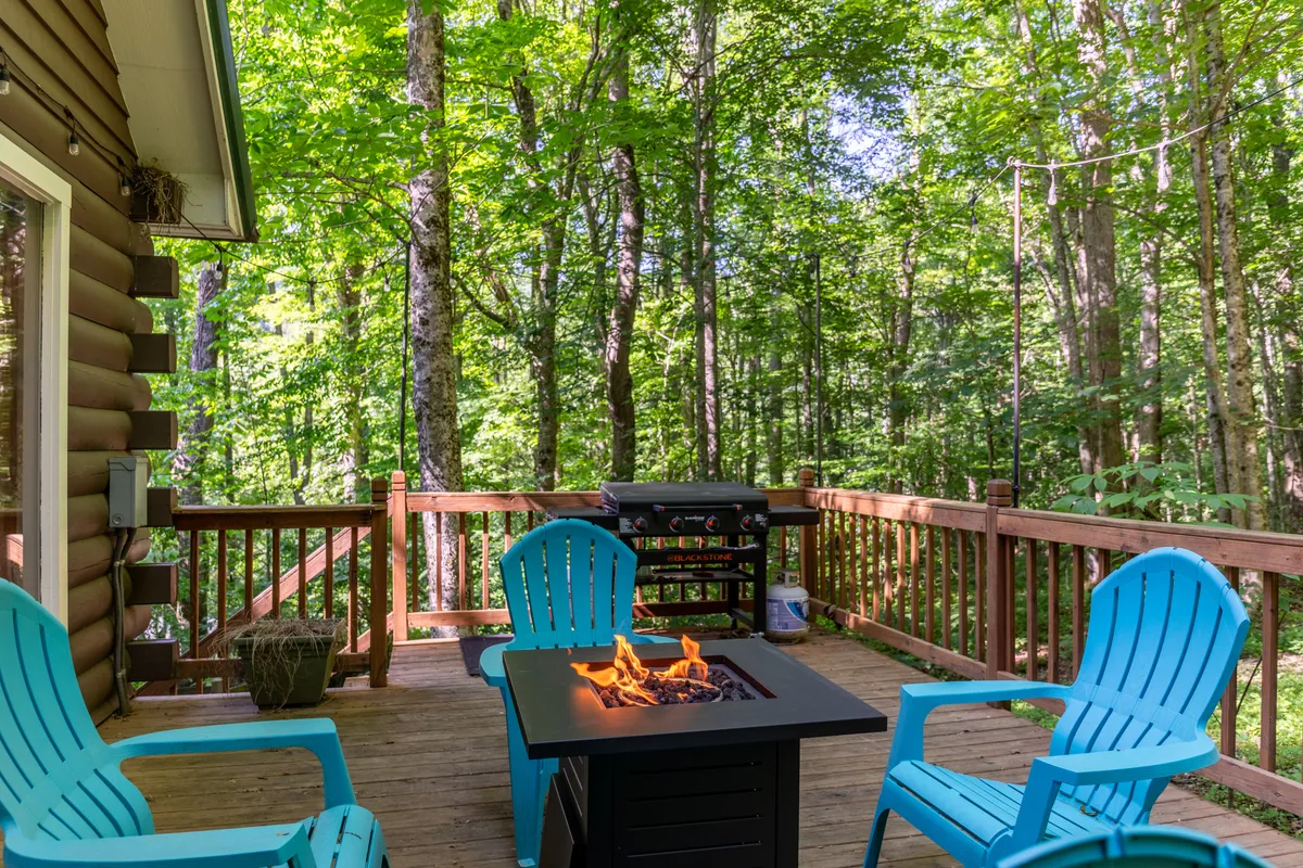 This image features a wooden deck surrounded by tall trees, with a fire pit at the center encircled by blue chairs and a grill nearby. The setting radiates a cozy ambiance, perfect for gatherings at sunset or under the stars.