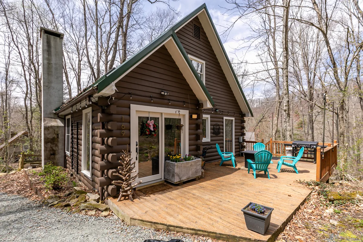 This image features a rustic log cabin structure with a spacious deck, comfortable seating, and large windows, surrounded by vibrant planters with flowers. The natural landscape around the cabin is lush and inviting, evoking the spirit of the High Country.