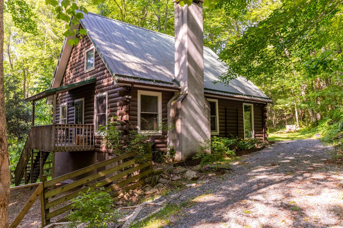 This exterior shot captures a charming log cabin nestled in lush greenery, with a gravel path leading to its covered porch and a chimney visible on the side. The cabin is surrounded by natural beauty, evoking the sense of seclusion and serenity ideal for a couples’ mountain retreat.