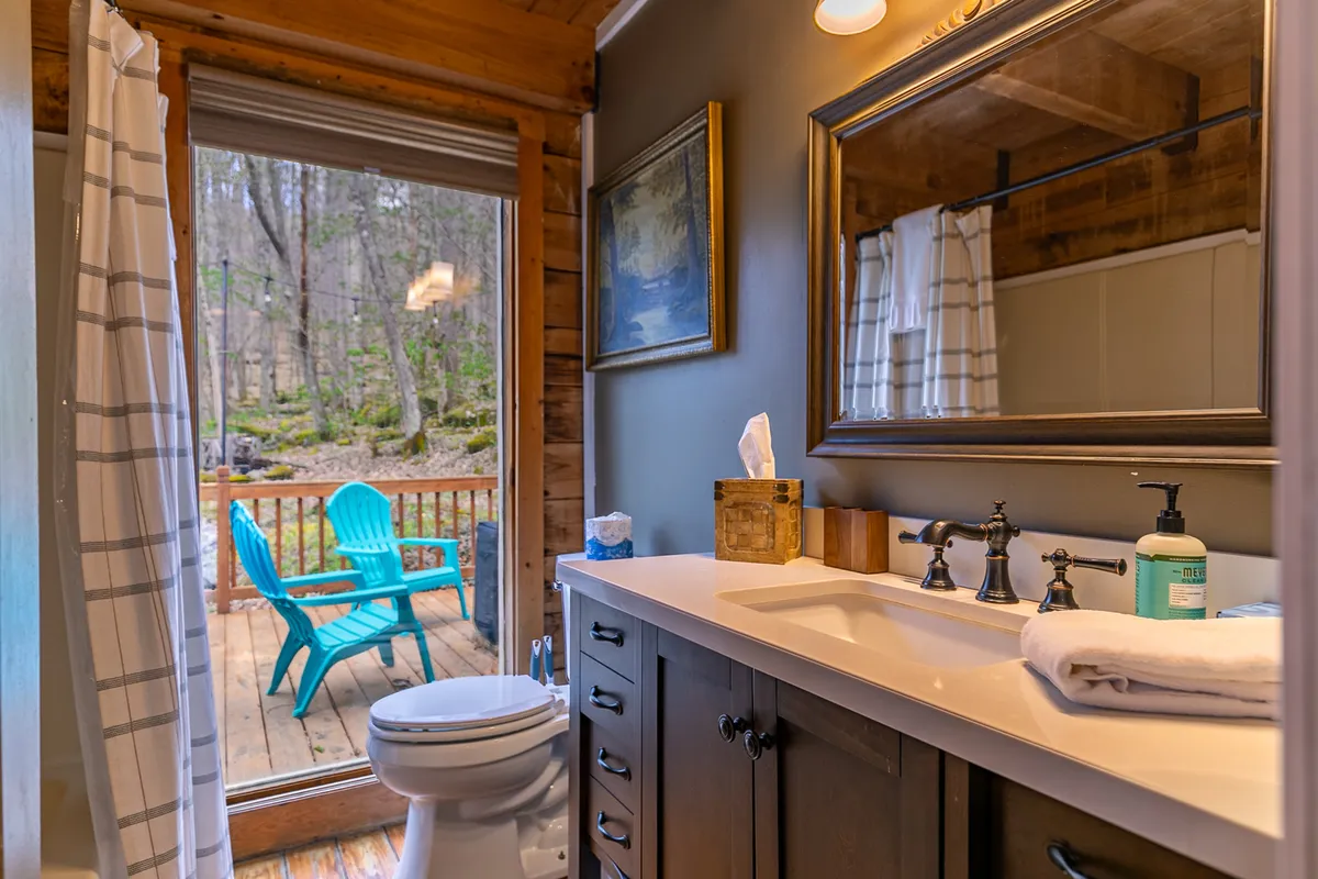 A tranquil bathroom scene with a vanity and wooden cabinetry, complemented by framed artwork and a sliding glass door leading to an outdoor deck. The door and deck offer a direct visual connection to nature, reinforcing the cabin’s integration with its historic surroundings.